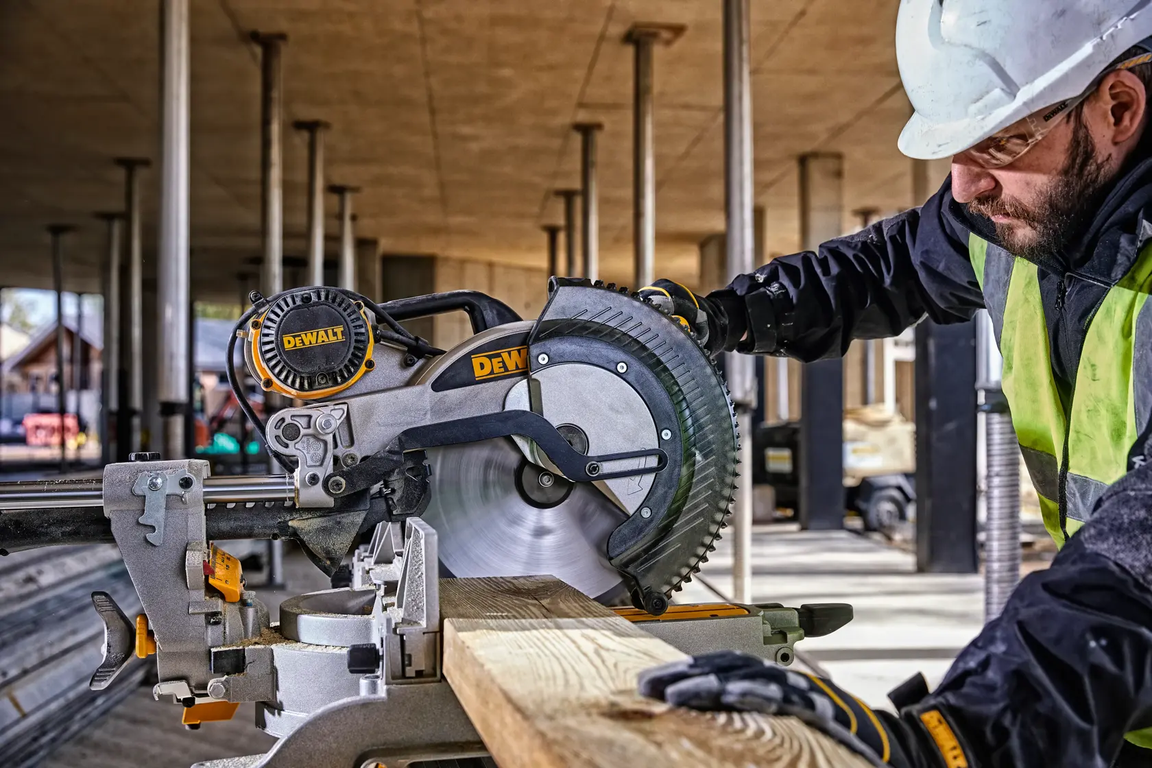 A person wearing a white safety helmet and gloves operates a yellow DEWALT DWS780 miter saw at a construction site. The background shows an unfinished building structure with columns and beams.