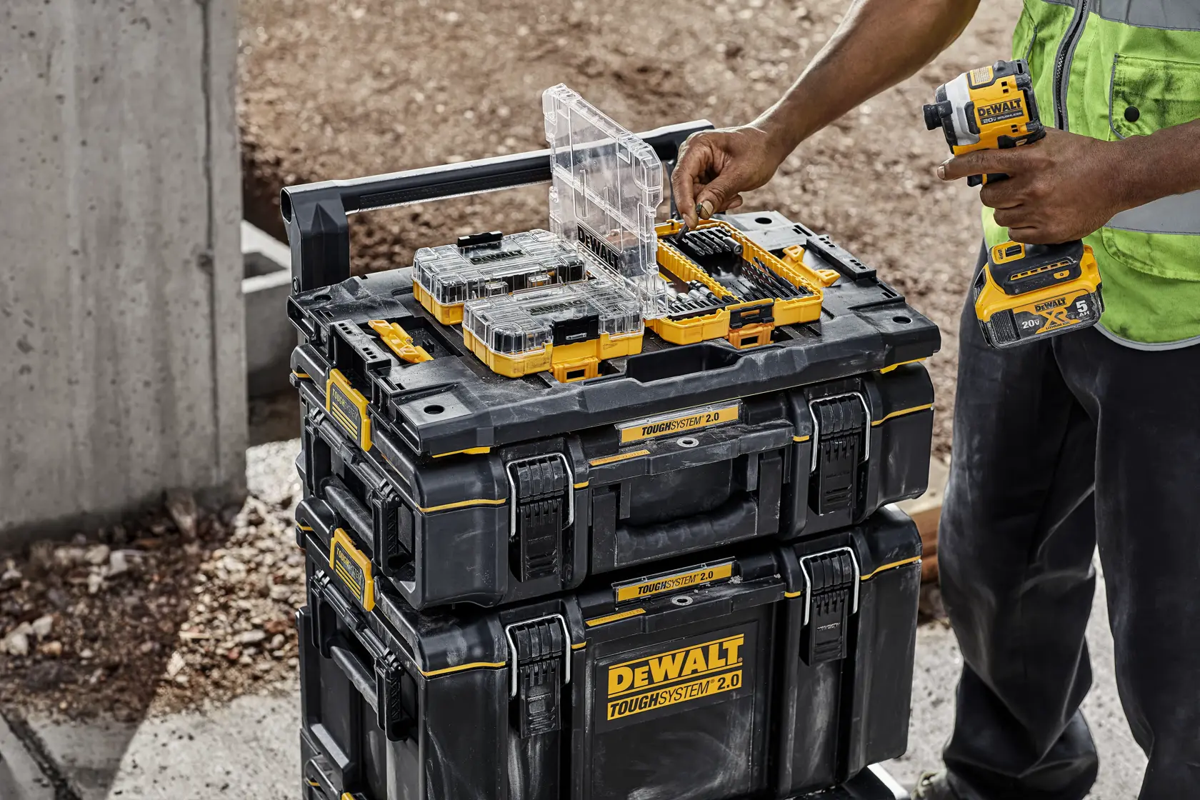A person on a construction site organizes drill bits and accessories on top of stacked DEWALT ToughSystem 2.0 toolboxes, using a DEWALT power tool.