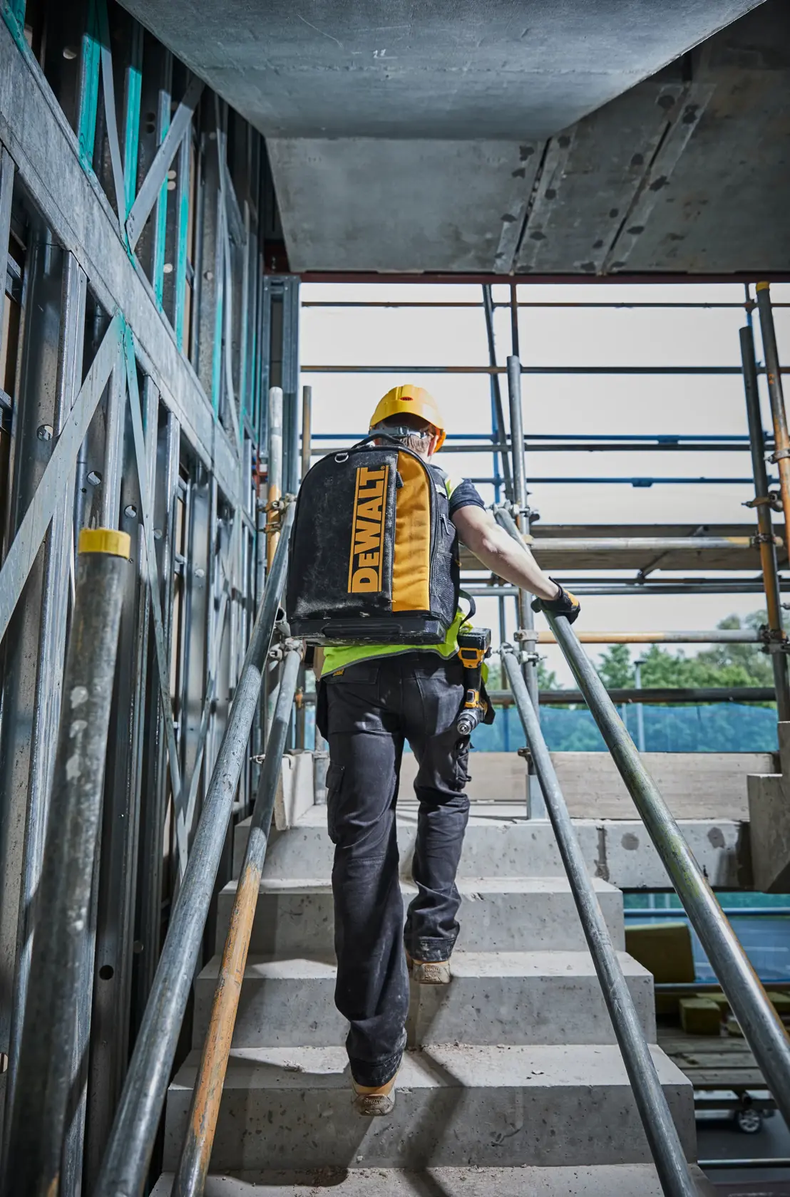 Operaio con zaino DEWALT su scale in cantiere, indossa casco giallo e attrezzatura di sicurezza.