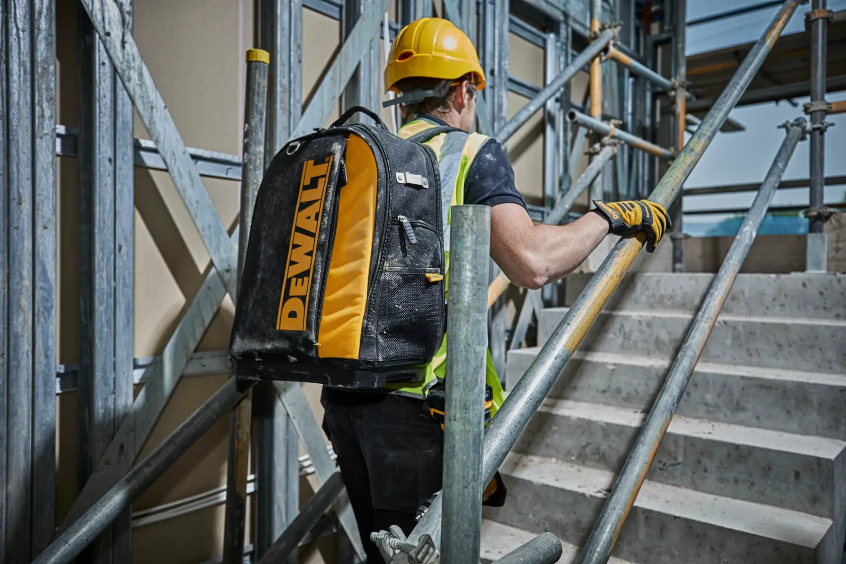 Operaio con zaino DEWALT sale le scale in cantiere, indossa casco e abbigliamento protettivo.