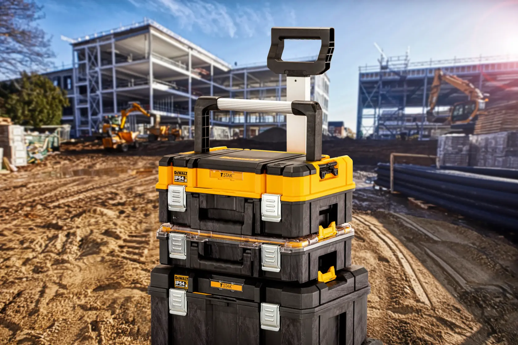 Cassette per attrezzi DEWALT impilate con manico su cantiere, strutture metalliche e macchine sullo sfondo.