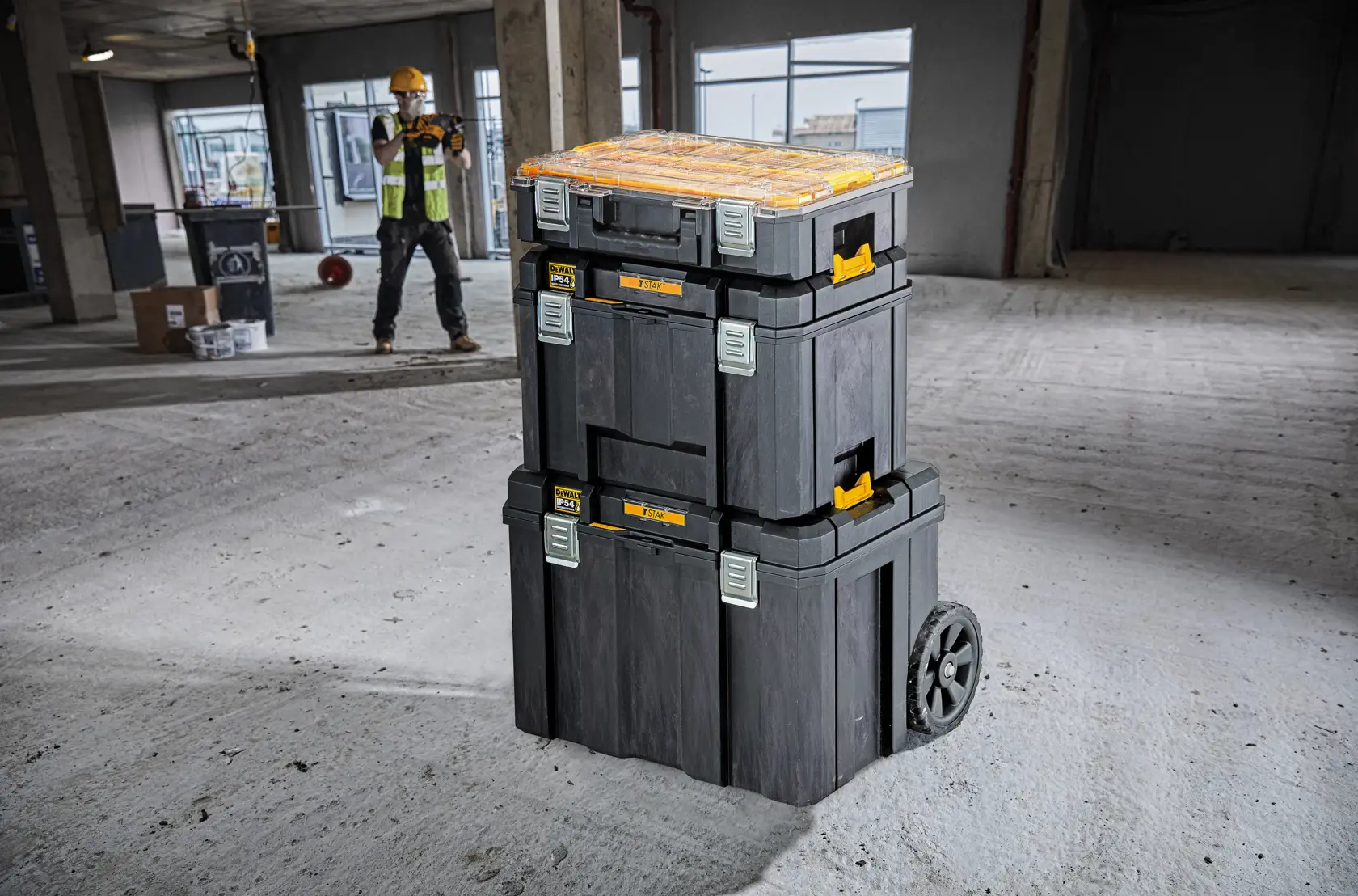 Cassette degli attrezzi DEWALT impilate con ruote su un cantiere, operaio sullo sfondo.